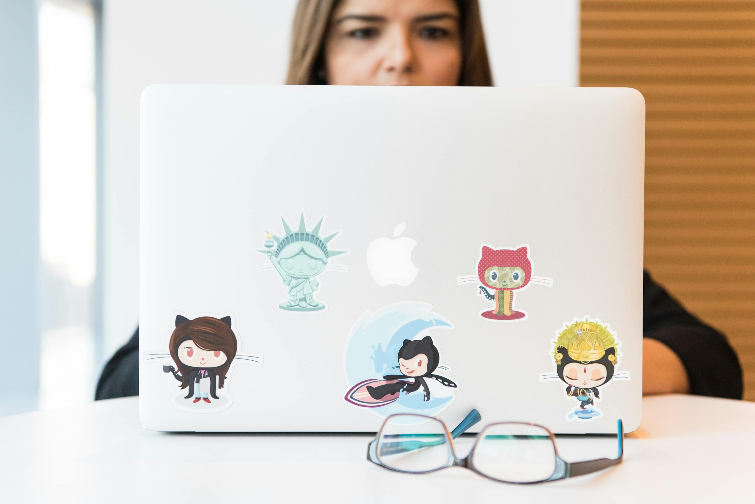 Woman focused on laptop with colorful GitHub-themed stickers, eyeglasses in foreground.